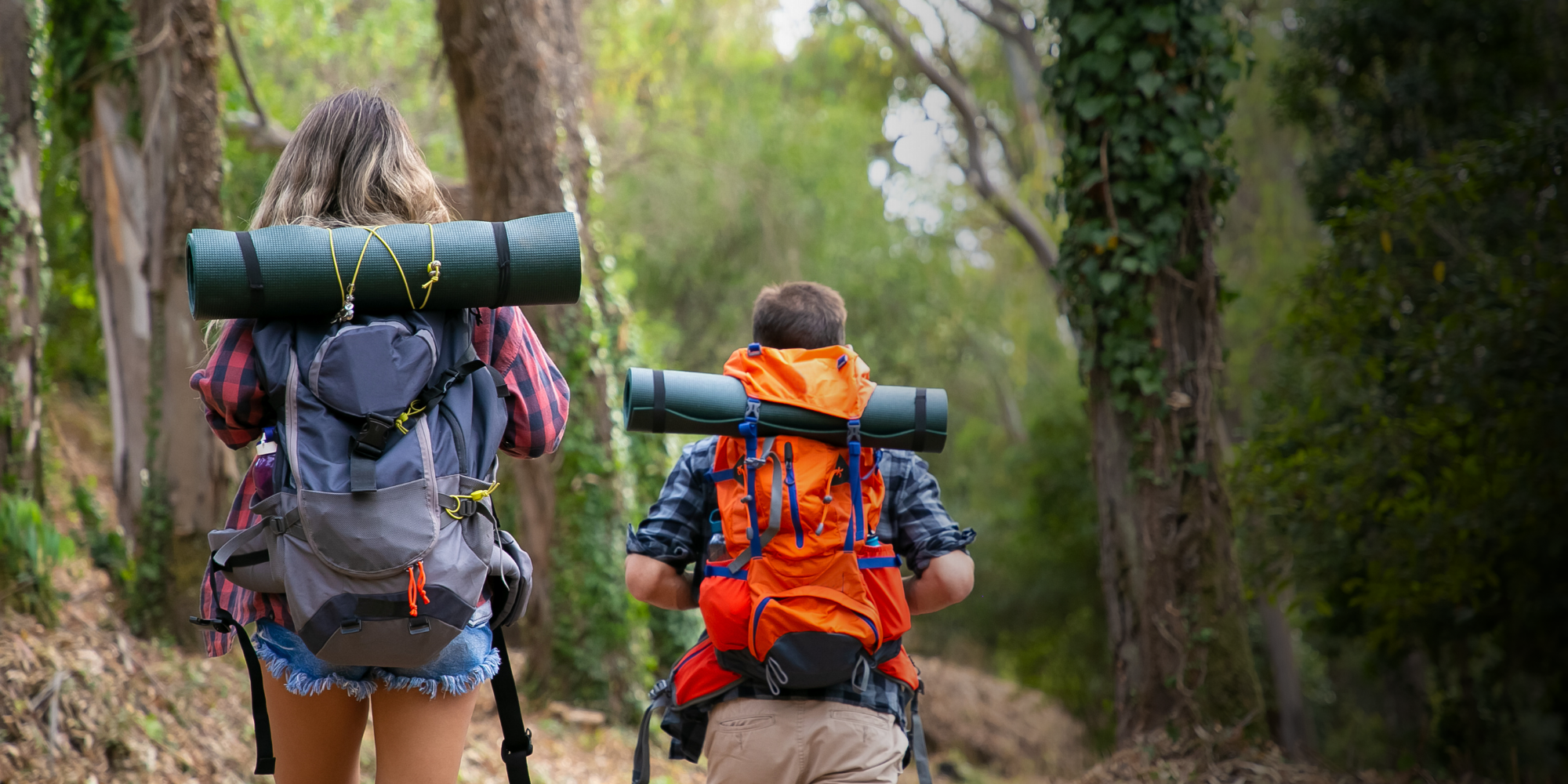 Two people hiking in a forest with backpacks and camping gear.

